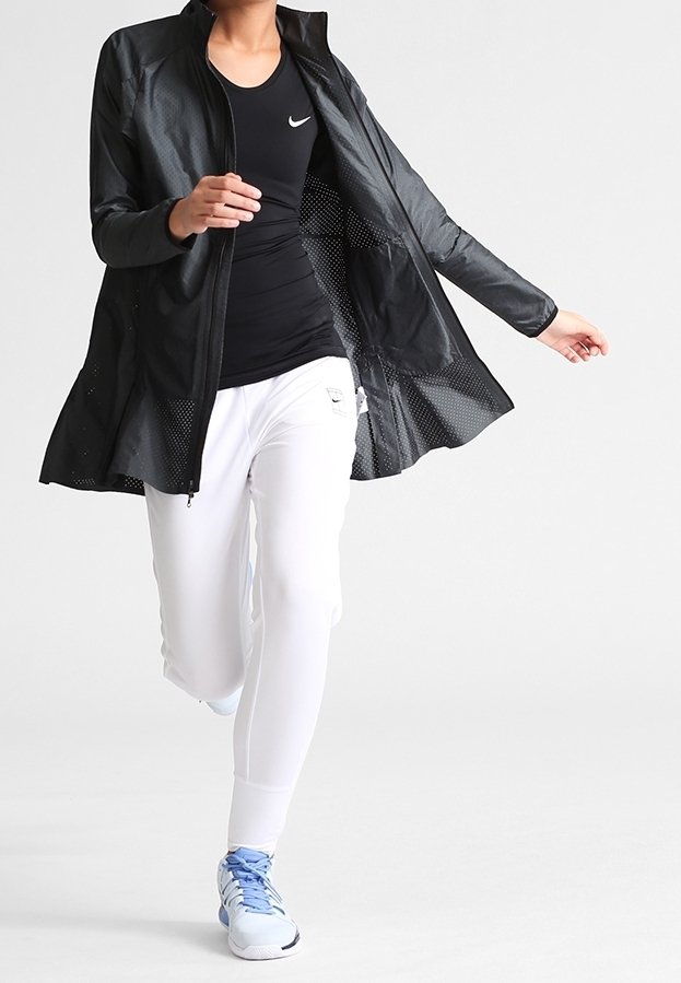 Chaqueta ligera negra con paneles de malla, combinada con una camiseta negra y pantalones ajustados blancos. El conjunto incluye zapatillas deportivas azules.