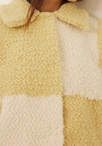 Puchaty, szlachetny płaszcz z naprzemiennymi kwadratami miękkiego żółtego i jasnobeżowego materiału o fakturowanej powierzchni oraz klasycznym kołnierzem.