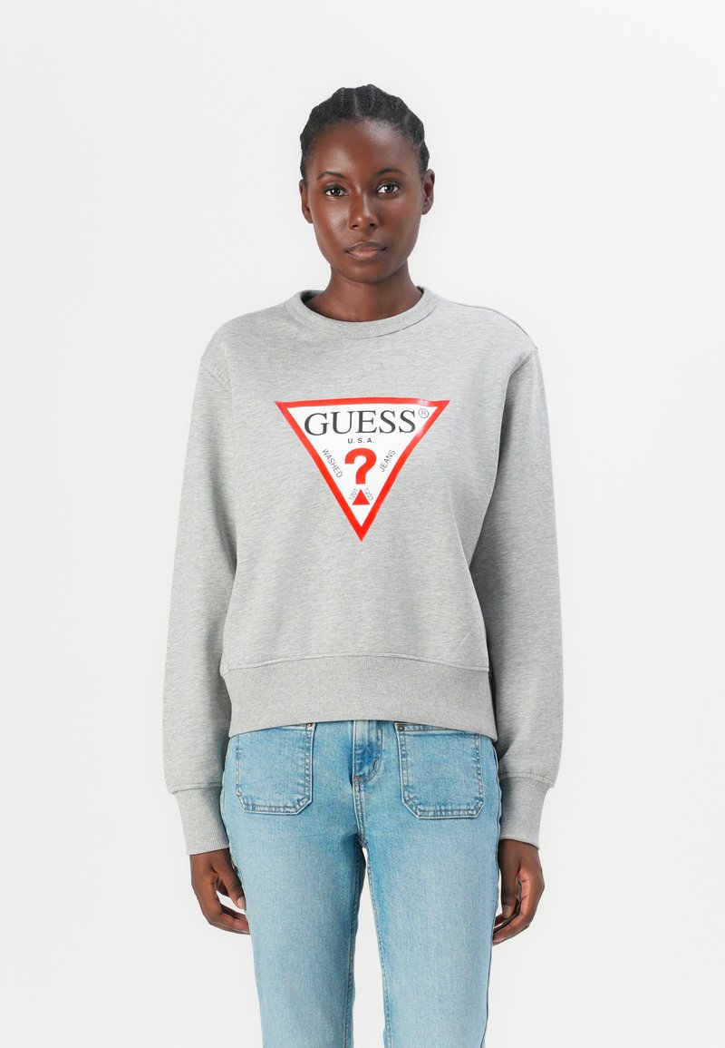 Grijze sweatshirt met een brede halslijn en geribde boorden, met een rood driehoekig logo met de tekst "GUESS?" in het midden. Gepaard met lichtblauwe jeans.