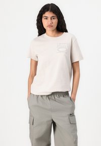 Šviesiai rudos spalvos medvilninis t-shirt trumpomis rankovėmis, su baltais spausdintais Nike logotipais. Suderintas su šviesiai pilkais krovinių kelniais su šoninėmis kišenėmis.