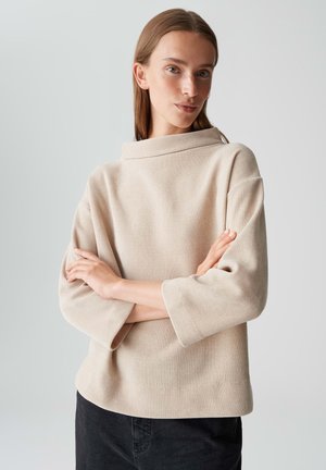 Jeune femme à la peau claire et aux cheveux raides, portant un pull beige texturé à manches larges et un pantalon noir, les bras croisés, devant un fond uni.