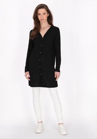 Abito nero a maniche lunghe con bottoni, scollatura a V, caratterizzato da bottoni marroni e una vestibilità comoda, abbinato a pantaloni bianchi e sneakers.