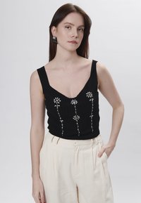 Top nero senza maniche con scollatura a V, tessuto testurizzato e ricami floreali con dettagli argentati. Abbinato a pantaloni color crema.