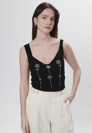 Zwarte mouwloze top met een V-hals, textuurstof en bloemenborduursel met zilveren accenten. Gepaard met crèmekleurige broeken.