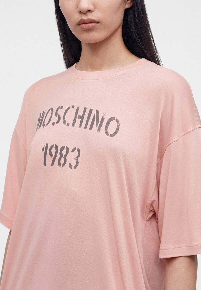 Woman wearing loose, pale pink T-shirt with black printed text "MOSCHINO 1983" across the chest, standing against a plain light background.