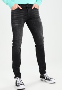 Mustad slim-fit teksad, millel on pleekinud tekstuur, viis taskut ja keskmise kõrgusega piht, koos mustade ja valgete spordijalanõudega.