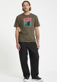 T-shirt en coton vert olive avec un design graphique coloré représentant une fleur et une figure. Coupe ample associée à un pantalon en denim foncé et des baskets noires.