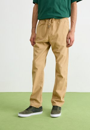 Beige cotton pants with an elastic waistband, front drawstring, and side pockets. Paired with green sneakers featuring a black logo.