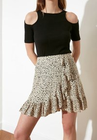 Zwarte cold-shoulder top gecombineerd met een beige rok met een zwart gespikkeld patroon, ruches aan de zoom en wikkelontwerp.