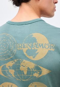 Κοντινό πλάνο ενός ατόμου που φορά πράσινο πουκάμισο με κίτρινα αφηρημένα σχέδια υδρογείου και το κείμενο «RUN AMOK» στην πλάτη.