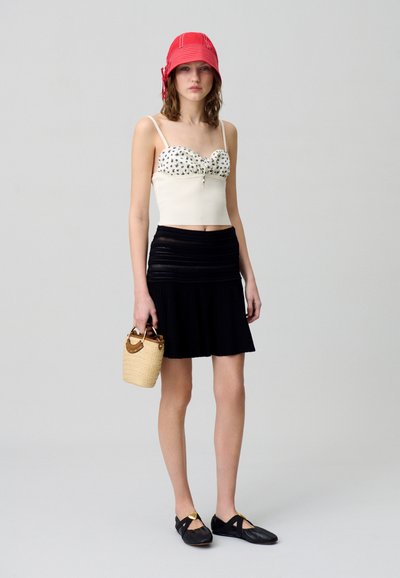 Cropped top bianco a pois con dettaglio a fiocco, abbinato a una gonna plissettata nera. Gli accessori includono un cappello a secchiello rosso e una piccola borsa intrecciata. Scarpe flat nere.
