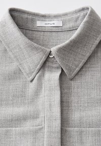 Camicia grigia con texture, colletto pieghevole, chiusura a bottoni e un'etichetta con la dicitura "OPUS". Il tessuto presenta un motivo sottile.