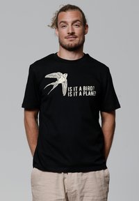 Zwarte katoenen t-shirt met een grafiek van een vliegende vogel en de tekst "Is het een vogel? Is het een vliegtuig?" in lichtgekleurde letters. Eenvoudig ontwerp.