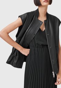 Black leather sleeveless jacket with ribbed texture and snap buttons, worn over a black pleated dress with a textured pattern.