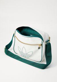 White synthetic bag with gold zipper, green accents, and perforated design. Features a large embroidered logo on the front and a adjustable strap.