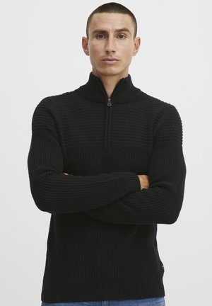 Schwarzer Strickpullover mit einem halbhohen Reißverschlusskragen. Strukturiertes Muster auf der Brust, glatte gerippte Ärmel und Saum, lockere Passform und Stil.