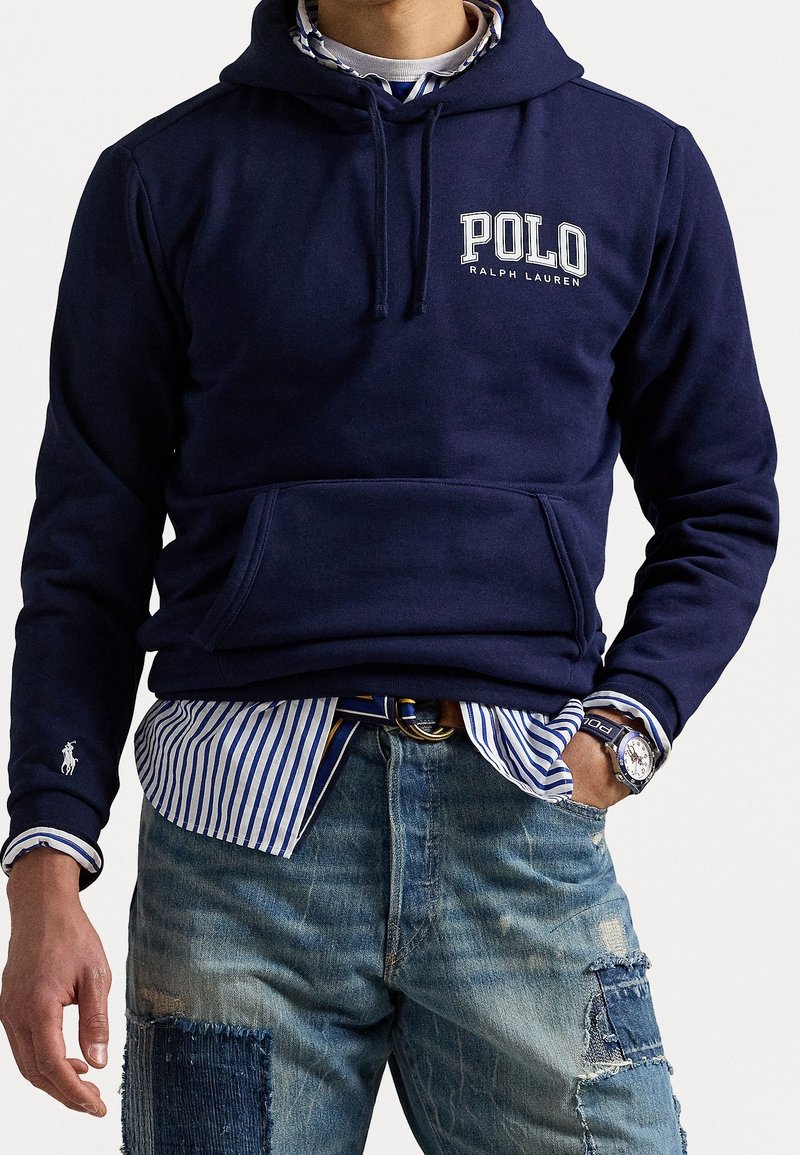 Námořnická mikina s logem "POLO", s přední kapsou a kapucí na šňůrku, vrstvená přes modro-bíle pruhovanou košili a potrhané modré džíny.