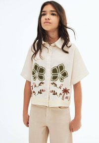 Blusa a maniche corte di colore crema con dettagli in pizzo all'orlo, presenta ricami di foglie verdi e motivi floreali, con chiusura a bottoni sul davanti.