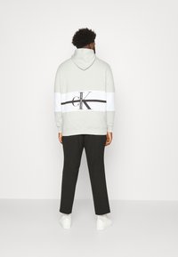 Hombre de espaldas con una sudadera ligera gris con una franja de logo de Calvin Klein en blanco y negro, pantalones negros, calcetines blancos y zapatillas blancas.