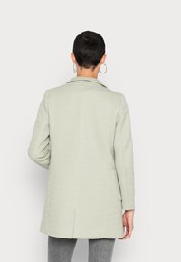 Jasnozielony płaszcz w wzór rybiej ości z wyciętym kołnierzem, długimi rękawami i bocznymi kieszeniami, zaprezentowany od tyłu.