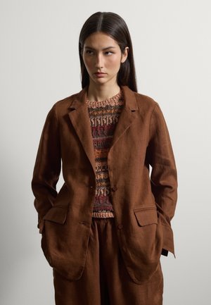 Junge Frau mit langen dunklen Haaren trägt einen lässigen braunen Blazer und Hose über einem mehrfarbigen Strickpullover und steht vor einem hellen, einfarbigen Hintergrund.