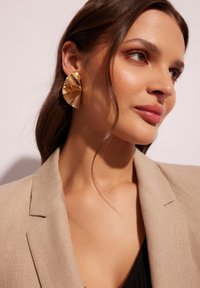 Złote kolczyki o wyrazistym designie, z rzeźbioną formą, charakteryzujące się złożonym kształtem i błyszczącą powierzchnią, noszone z beżową marynarką.
