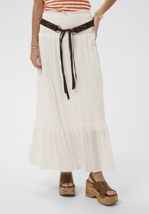 Femme portant une longue jupe blanche avec une taille smockée, une ceinture marron nouée, un haut rayé orange, des sandales compensées marron et des bracelets dorés.