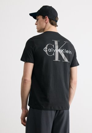 Mand iført sort kasket og sort Calvin Klein t-shirt med stort "cK" logo på ryggen, stående foran en ensartet hvid baggrund.