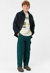 Paitapaita, jossa on tummanvihreä ja laivastonsininen ruutukuosi, päällä vaalea graafinen t-paita. Tummanvihreät cargo-housut suurilla sivutaskuilla ja ruskeat saappaat.