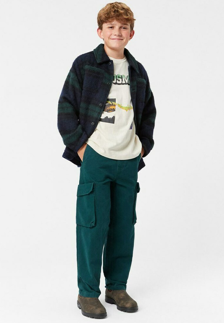 Paitapaita, jossa on tummanvihreä ja laivastonsininen ruutukuosi, päällä vaalea graafinen t-paita. Tummanvihreät cargo-housut suurilla sivutaskuilla ja ruskeat saappaat.