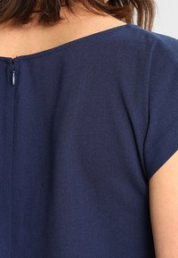 Haut bleu marine avec un décolleté bateau et des manches courtes, présentant une fermeture éclair arrière visible. Tissu texturé avec un éclat subtil.