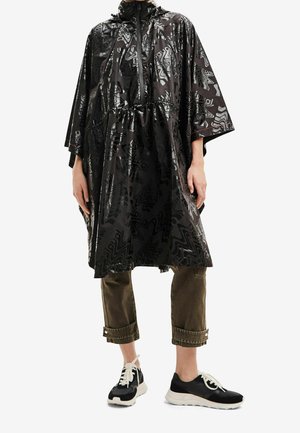 Personne portant un poncho de pluie noir à motifs avec une fermeture éclair et une taille à cordon, un pantalon marron raccourci et des baskets noires et blanches.