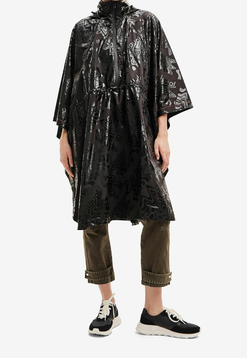 Personne portant un poncho de pluie noir à motifs avec une fermeture éclair et une taille à cordon, un pantalon marron raccourci et des baskets noires et blanches.