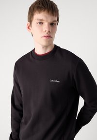 Crna dukserica izrađena od glatkog materijala, s okruglim izrezom i malim bijelim logotipom "Calvin Klein" na lijevoj strani prsnog koša.