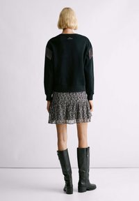 Felpa nera con dettagli a frange, indossata sopra una gonna mini fluida in bianco e nero con motivo. Indossatore in stivali neri con tacco alto.