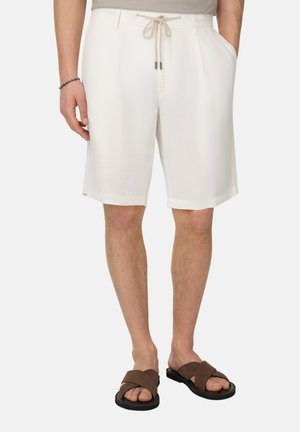 Man die witte short met trekkoord en bruine sandalen met kruisbandjes draagt, staat met handen in de zakken tegen een effen achtergrond.