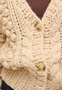 Dzianinowy kardigan w jasnym beżu z teksturowanym wzorem, z dużymi, wyraźnymi kablami i dwoma okrągłymi guzikami z przodu.