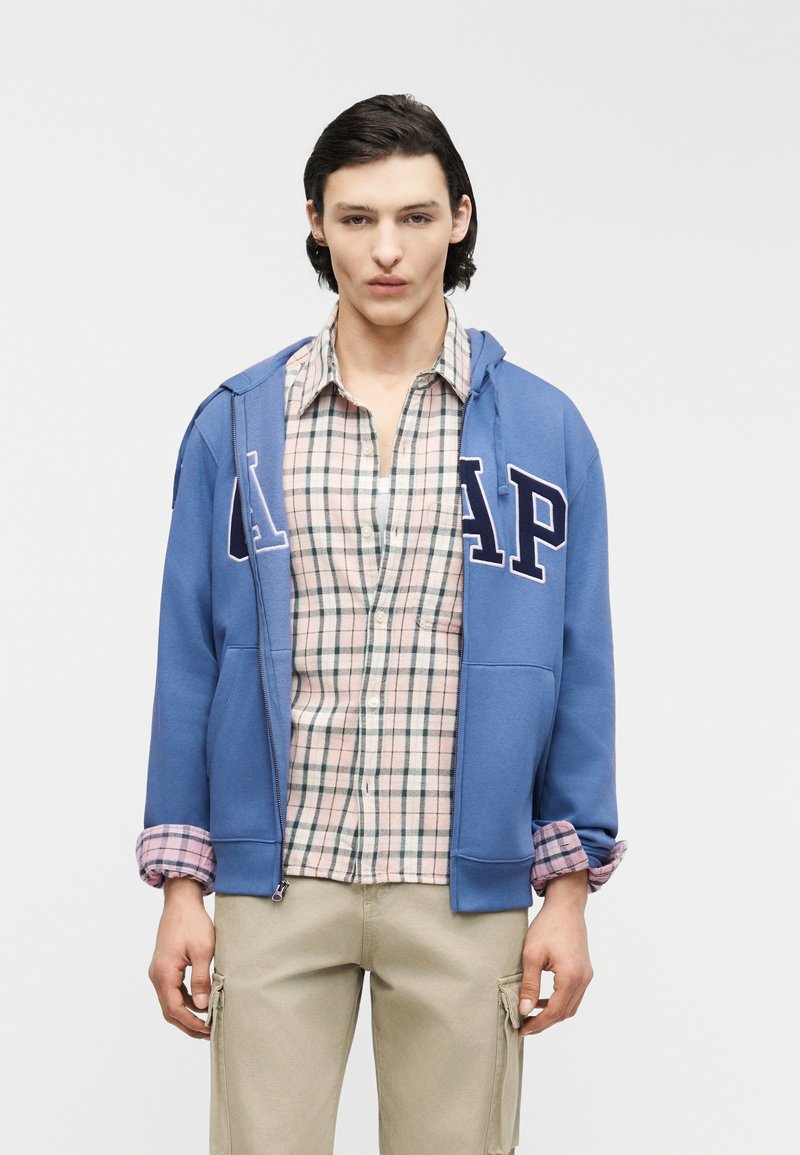 Jeune homme portant un sweat à capuche bleu de la marque GAP par-dessus une chemise à carreaux rose et verte à boutons et un pantalon cargo beige, debout devant un fond uni.