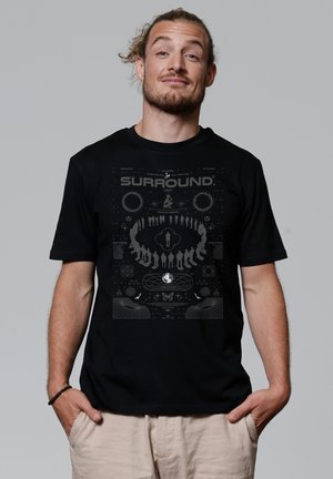 Zwarte katoenen t-shirt met een gedurfde grafische print in grijs met de tekst "SURROUND," abstracte vormen en figuren gerangschikt in een cirkelontwerp.