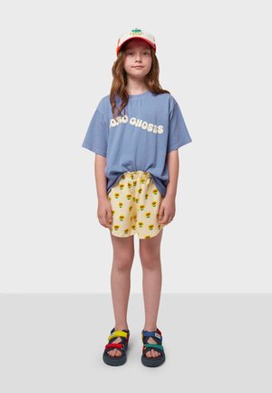 T-shirt azzurro a maniche corte con il testo "100% CHOSES", pantaloni corti gialli con stampa di girasoli verdi e neri, e sandali multicolori.