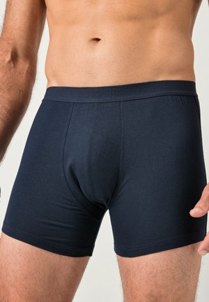 Pánský trup a horní část stehen v jednobarevných tmavě modrých přiléhavých boxerkách na neutrálním pozadí.