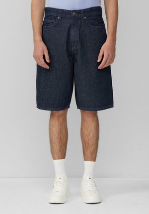 Shorts en denim foncé avec une coupe droite, un design à cinq poches et une fermeture à bouton, associés à des baskets blanches et des chaussettes chevilles.