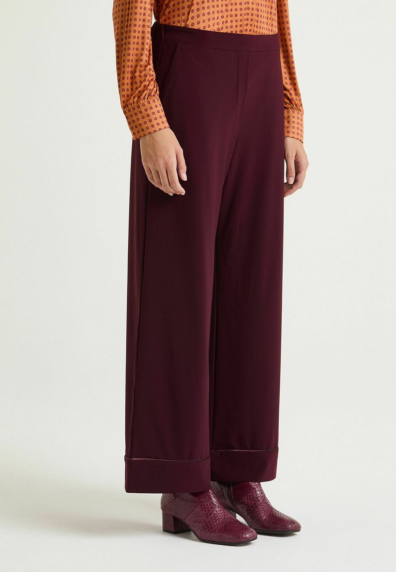 Pantaloni bordeaux a gamba larga con una texture liscia, dotati di una vita sagomata e risvolti arrotolati, abbinati a stivaletti bordeaux lucidi.