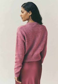 Kvinna med mörkt hår som bär guldörhängen, en rosa stickad tröja och en matchande rosa kjol, sedd från ryggen och sidan.
