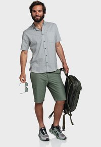 Hellgraues kurzärmliges Hemd, mit Knopfleiste und Tasche, kombiniert mit grünen Shorts. Mann hält eine Sonnenbrille, trägt einen dunkelgrünen Rucksack und graue Sneakers.