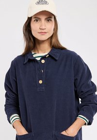 Chemise en velours côtelé bleu marine avec un col boutonné, featuring des boutons en bois et deux poches avant. Superposée avec une chemise rayée en dessous.