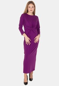 Vestito viola a maniche lunghe con design attorcigliato sul davanti, silhouette aderente e tessuto liscio, abbinato a tacchi alti neri.