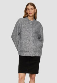 Graue Wolljacke mit strukturierter Oberfläche, hohem Kragen, Fronttaschen und einem leicht oversized Schnitt, getragen über einem schwarzen Rock.