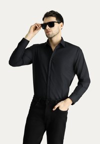 Camicia nera a maniche lunghe con chiusura a bottoni e taschino sul petto, indossata con occhiali da sole neri e jeans neri, su uno sfondo neutro.