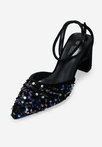 Zapato de tacón de lentejuelas negras con punta afilada y tacón ancho, con finas cintas negras alrededor del tobillo. Lentejuelas brillantes de varios colores cubren la parte superior.